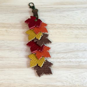 May include: A fall-themed keychain featuring a cascade of leather maple leaves in shades of red, orange, yellow, and brown. The leaves are attached to a brown leather strap and a bronze-colored clasp.