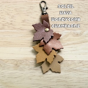 May include: A leather keychain featuring four layered maple leaf cutouts in shades of dark brown, medium brown, tan, and beige, attached to a bronze-toned metal lobster clasp. The text SOLEIL HAVA HONEYCOMB CHAMPAGNE is printed in white.
