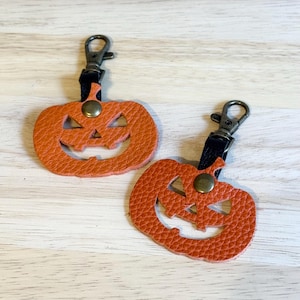 May include: Two orange leather pumpkin keychains with carved faces and metal clasps. The keychains have a textured surface and black straps. The clasps are attached to the top of the pumpkins.
