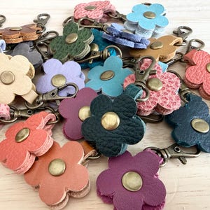 May include: A collection of flower-shaped keychains in various colors, including pink, blue, green, and purple. Each keychain features a metal clasp and a central brass-colored button. The keychains are made of leather.