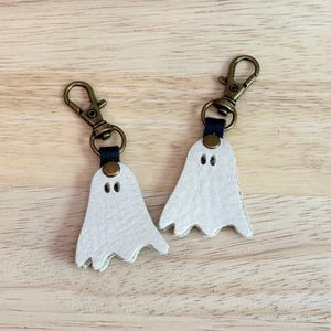 May include: Two white leather ghost keychains with bronze-colored clasps and black leather accents. Each ghost has two small eye holes and a jagged bottom edge. The keychains are displayed on a light brown wooden surface.