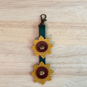 May include: A dark green leather strap keychain featuring two layered sunflower-shaped charms. Each flower has yellow textured petals and a dark red circular center with a metallic brass-toned snap button. A bronze-colored lobster clasp is attached.