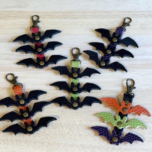 May include: Five leather bat keychains with bronze-colored hardware. Each bat is black with colorful accents in orange, green, purple, pink, and purple. The keychains are arranged on a light wood surface.