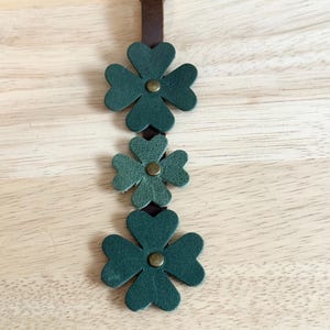 May include: A dark green leather keychain featuring three four-leaf clovers. The clovers are attached to a brown leather strap with a bronze-colored clasp. The keychain is a symbol of good luck.
