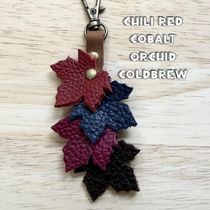 May include: A leather keychain featuring four layered maple leaf cutouts in chili red, cobalt, orchid, and coldbrew colors. The leaves are attached to a brown leather strap with a metal swivel clasp and a gold-toned rivet at the top.