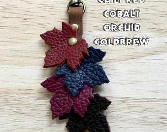 May include: A leather keychain featuring four layered maple leaf cutouts in chili red, cobalt, orchid, and coldbrew colors. The leaves are attached to a brown leather strap with a metal swivel clasp and a gold-toned rivet at the top.