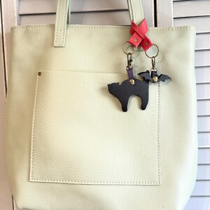 May include: A light green leather tote bag with a large front pocket. Attached to the handle is a pink leather accent with two keychains: a black cat and a black bat. The bag has a simple, minimalist design.