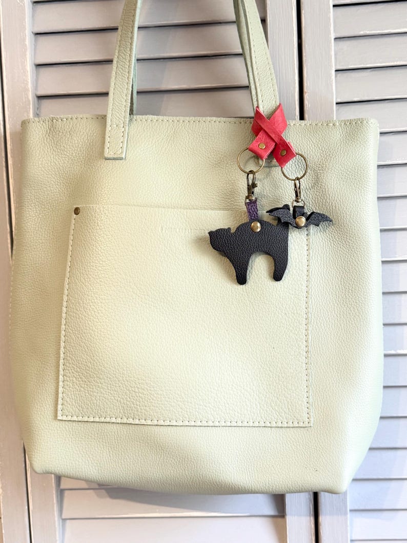 May include: A light green leather tote bag with a large front pocket. Attached to the handle is a pink leather accent with two keychains: a black cat and a black bat. The bag has a simple, minimalist design.