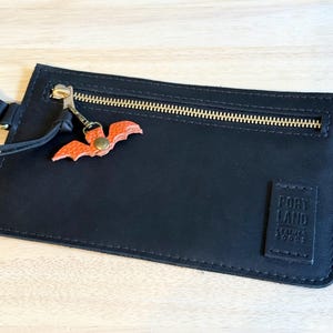 May include: Black leather wallet with a gold zipper and a small orange bat charm. The wallet has a rectangular shape and a tag that reads "PORTLAND LEATHER GOODS". The bat charm is attached to the zipper pull.
