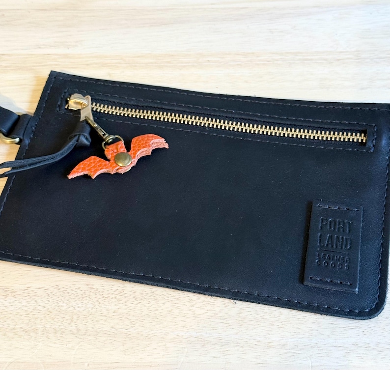 May include: Black leather wallet with a gold zipper and a small orange bat charm. The wallet has a rectangular shape and a tag that reads "PORTLAND LEATHER GOODS". The bat charm is attached to the zipper pull.