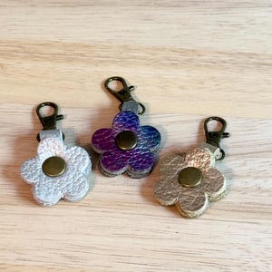 May include: Three flower-shaped keychains in silver, iridescent purple, and gold. Each keychain has a metal clasp and a central button. The keychains are made of a textured material and are displayed on a light wooden surface.
