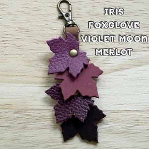 May include: A leather keychain featuring four layered maple leaf shapes in shades of purple and dark red, attached to a bronze-toned metal lobster clasp. The text IRIS, FOXGLOVE, VIOLET MOON, and MERLOT is printed in white on the right side.