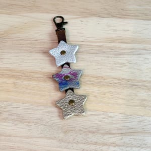 May include: A leather keychain with three star-shaped charms. The top and bottom stars are silver, and the middle star has a colorful, tie-dye effect. The keychain has a brown leather strap and a bronze-colored clasp.