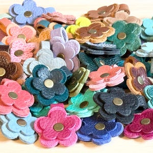 May include: A collection of colorful leather flower-shaped accessories. The flowers are in various shades of pink, blue, green, brown, and purple, each with a brass-colored center. Some have loops attached.