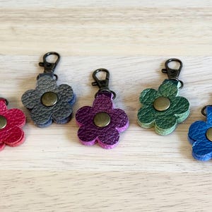 May include: Five leather flower-shaped keychains in shades of red, gray, purple, green, and blue. Each keychain has a metal clasp and a central button. The keychains are arranged on a light wood surface.