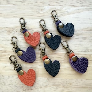 May include: A collection of six heart-shaped keychains made from textured material in orange, purple, and black. Each keychain features a bronze-toned metal lobster clasp and a matching circular metal snap button fastener at the top center.