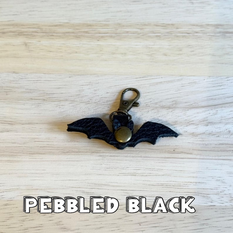 May include: A black, bat-shaped keychain made of pebbled leather. The keychain has a bronze-colored clasp and a circular rivet. The text "PEBBLED BLACK" is displayed at the bottom of the image.