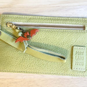 May include: A light green leather zippered pouch with a wrist strap and a small orange bat-shaped charm attached to the zipper pull. The pouch features a rectangular leather patch embossed with the text PORTLAND LEATHER GOODS.