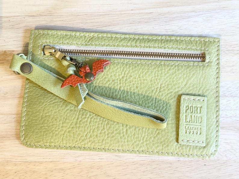 May include: A light green leather zippered pouch with a wrist strap and a small orange bat-shaped charm attached to the zipper pull. The pouch features a rectangular leather patch embossed with the text PORTLAND LEATHER GOODS.