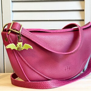 May include: A magenta-colored, crescent-shaped crossbody bag with a textured leather appearance. The bag features a long, adjustable strap and a small, green bat-shaped charm. The bag has a metal buckle and a stitched design.