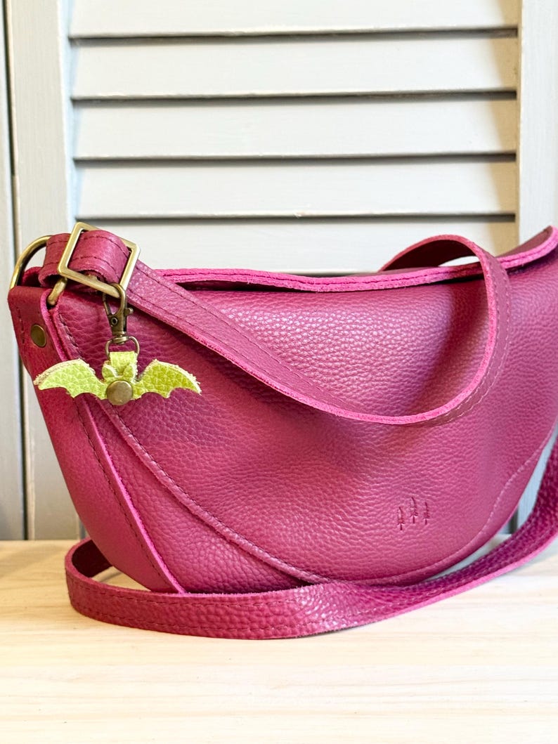 May include: A magenta-colored, crescent-shaped crossbody bag with a textured leather appearance. The bag features a long, adjustable strap and a small, green bat-shaped charm. The bag has a metal buckle and a stitched design.