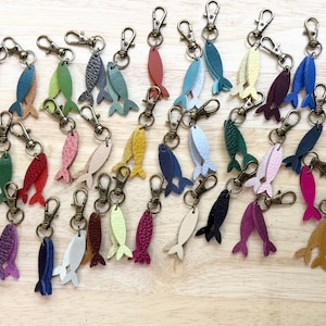 May include: A collection of numerous fish-shaped keychains made from various colored leather pieces. Each keychain features a metal lobster clasp attached to a small, flat, textured fish cutout. The assortment includes red, blue, green, yellow, and purple.