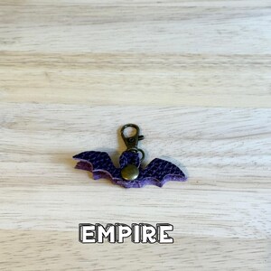 May include: A purple bat-shaped keychain made of textured material with a bronze-colored metal lobster clasp and a central circular snap button. The word EMPIRE is printed in bold white letters with a black outline below the bat.