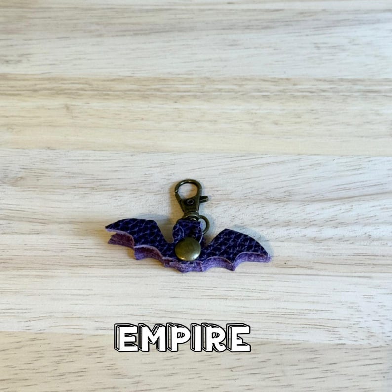 May include: A purple bat-shaped keychain made of textured material with a bronze-colored metal lobster clasp and a central circular snap button. The word EMPIRE is printed in bold white letters with a black outline below the bat.