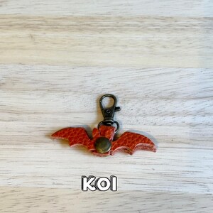 May include: A red leather bat-shaped keychain with a bronze-colored clasp and rivet. The bat has textured wings and the word "KOI" is printed at the bottom. The keychain is on a light wood grain surface.
