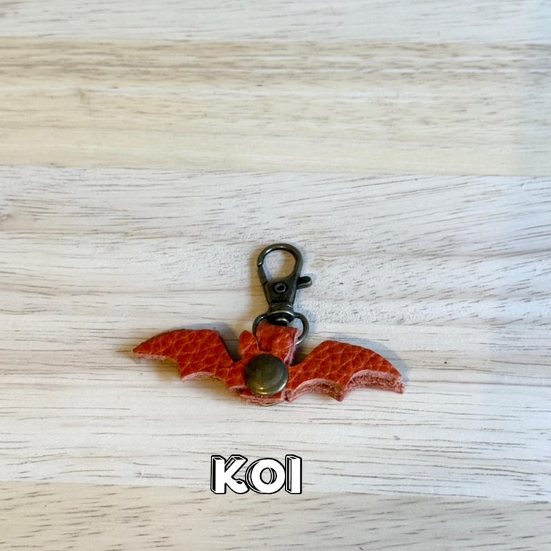 May include: A red leather bat-shaped keychain with a bronze-colored clasp and rivet. The bat has textured wings and the word "KOI" is printed at the bottom. The keychain is on a light wood grain surface.
