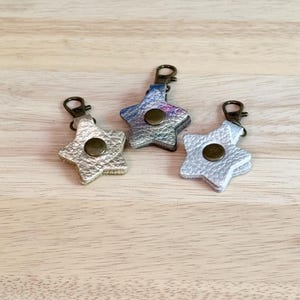 May include: Three star-shaped keychains in metallic silver, blue, and gold. Each keychain has a bronze-colored snap and a bronze-colored clasp. The keychains are displayed on a light brown wooden surface.