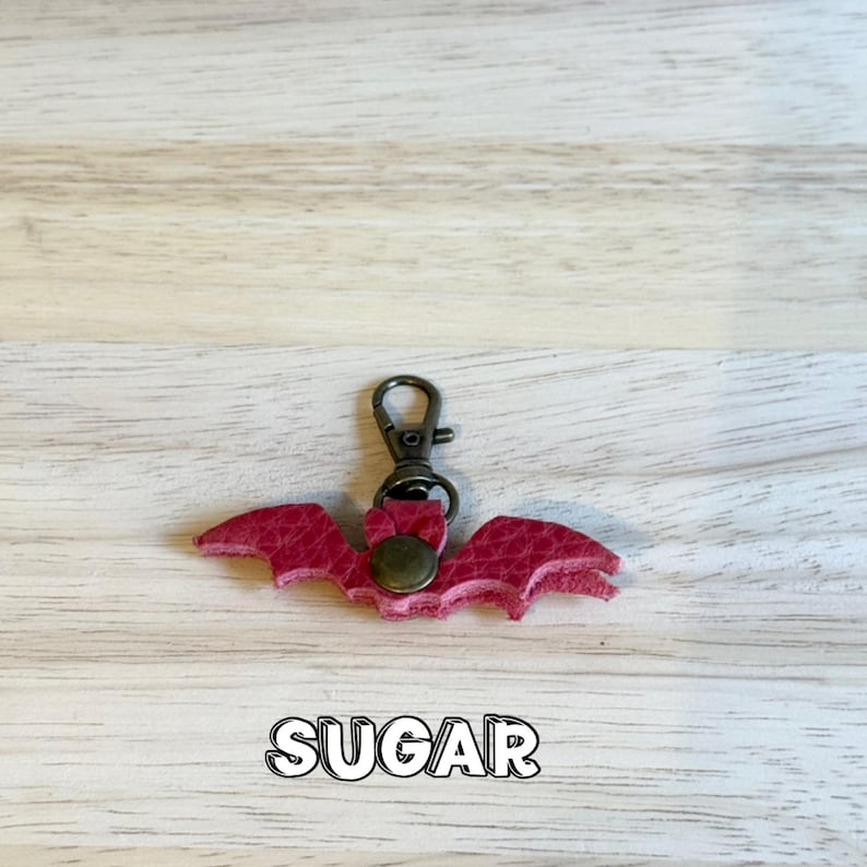 May include: A red bat-shaped keychain with a bronze-colored clasp and rivet. The word "SUGAR" is printed in a white, outlined font below the keychain. The keychain is made of textured material.