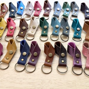 May include: A collection of numerous leather keychains in a wide variety of colors, each featuring a folded loop design secured with a metallic snap button and attached to a circular metal split ring for holding keys or other small items.