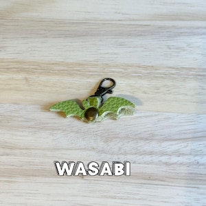 May include: A green leather bat-shaped keychain with a black clasp and a bronze-colored rivet. The word "WASABI" is printed in a white outlined font below the keychain. The keychain is on a light wood grain surface.