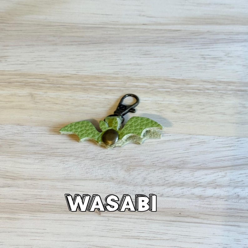 May include: A green leather bat-shaped keychain with a black clasp and a bronze-colored rivet. The word "WASABI" is printed in a white outlined font below the keychain. The keychain is on a light wood grain surface.