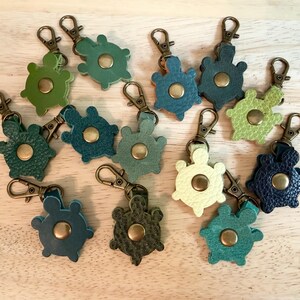 May include: A collection of turtle-shaped keychains crafted from textured leather in various shades of green, blue, and cream. Each keychain features a central brass-colored metal snap button and is attached to a brass-colored metal lobster clasp.