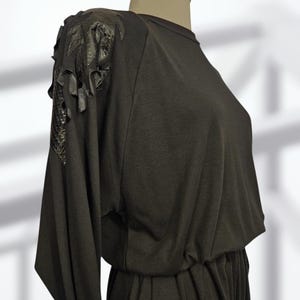 May include: A black dress with long sleeves and a gathered waist. The shoulders are embellished with decorative black leather or faux leather accents. The dress is displayed on a mannequin against a soft, neutral background.