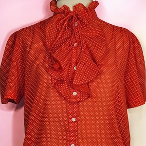May include: Red short-sleeved blouse with white polka dots. The blouse features a high ruffled collar and a cascading ruffle down the front, secured with a tie and buttons. The fabric appears lightweight and semi-sheer, creating a vintage aesthetic.