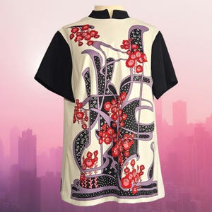 May include: Cream-colored tunic with black sleeves and collar. The front features a floral design in red, purple, and black. The tunic has a high collar and short sleeves, with a cityscape in the background.