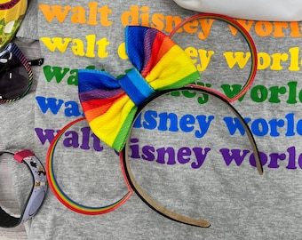 Op de afbeelding: Een grijs T-shirt met de tekst "walt disney world" die in verschillende kleuren wordt herhaald. Een regenboog hoofdband met een strik ligt op het shirt.