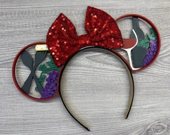 May include: A black headband with red sequin bow and two circular accents. One accent features crossed utensils, grapes, and greenery. The other shows a wine bottle, glass, and grapes. The accents are red with black and purple details.