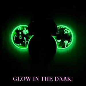 May include: Glow-in-the-dark decorative ears with a neon green outline. The ears feature intricate designs, including floral and skull motifs, illuminated against a dark background. The text "GLOW IN THE DARK!" is displayed in pink.