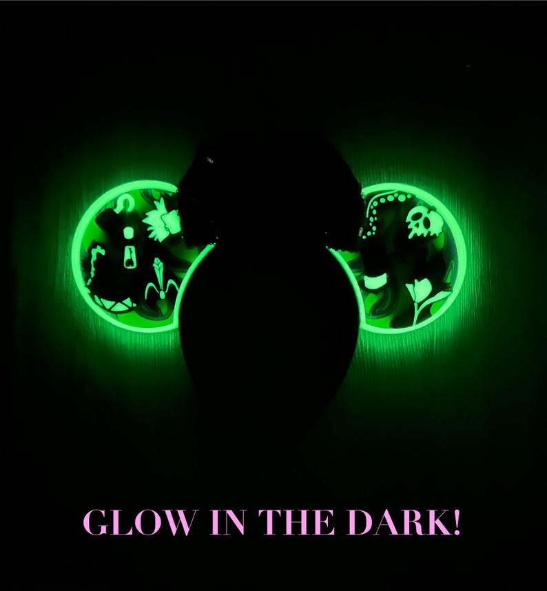 May include: Glow-in-the-dark decorative ears with a neon green outline. The ears feature intricate designs, including floral and skull motifs, illuminated against a dark background. The text "GLOW IN THE DARK!" is displayed in pink.