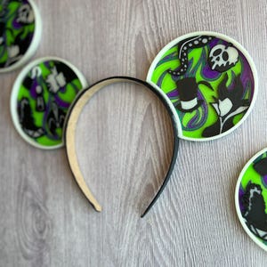May include: A black headband with decorative ear accents. The ears feature a green, purple, and black design with a skull, top hat, and other villainous elements. The ears are outlined in white.