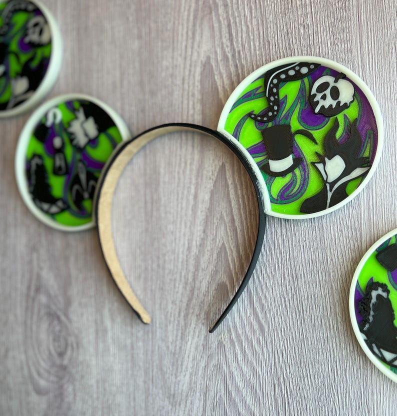 May include: A black headband with decorative ear accents. The ears feature a green, purple, and black design with a skull, top hat, and other villainous elements. The ears are outlined in white.