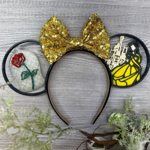 May include: Black mouse ears with a gold glitter bow, a red rose with green stem and leaves, and a white silhouette of Belle and the Beast in a yellow castle.