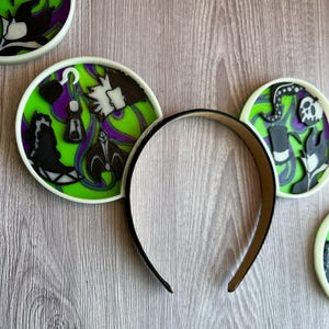 May include: A black headband with decorative round ear accents. The ear accents feature a lime green background with purple and black character designs. The designs include a hook, a bottle, and a skull.