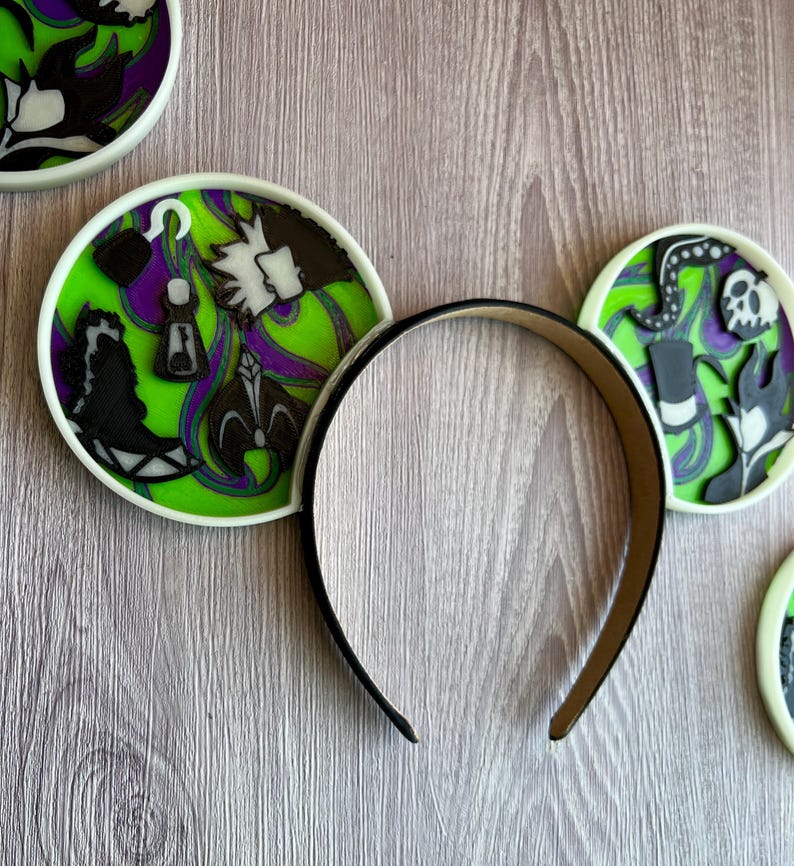 May include: A black headband with decorative round ear accents. The ear accents feature a lime green background with purple and black character designs. The designs include a hook, a bottle, and a skull.