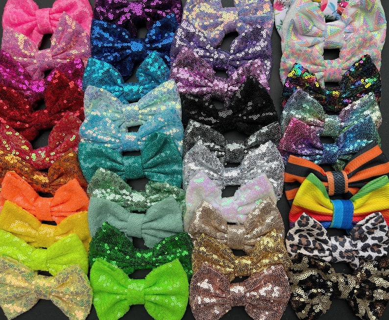May include: A collection of sequined hair bows in various colors, including pink, blue, green, and gold. The bows are arranged closely together, showcasing their sparkly texture and diverse hues. The bows are a mix of solid colors and patterned designs.