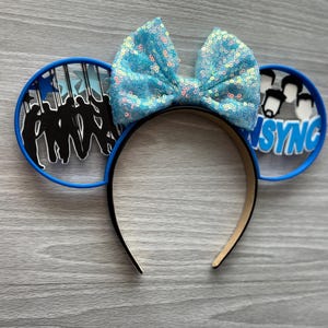 May include: A blue sequined bow headband with black and blue accents. The headband features two circular ear designs, one with a silhouette of a group of people and the other with the text "NSYNC".